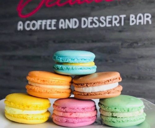 A colorful stack of macarons in various flavors sits on a plate, with a coffee and dessert bar sign in the background.