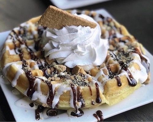 A delicious waffle topped with whipped cream, chocolate drizzle, crushed cookies, and a graham cracker.