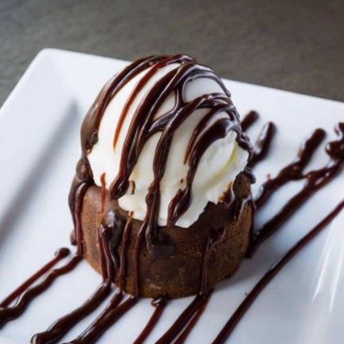 A warm chocolate cake topped with a scoop of vanilla ice cream and drizzled with chocolate sauce on a white plate.