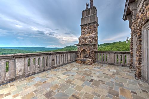 A stone terrace with a fireplace overlooks a scenic view of rolling hills under a cloudy sky.