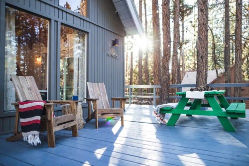 A cozy deck with two wooden chairs and a green picnic table, surrounded by tall trees and sunlight filtering through.