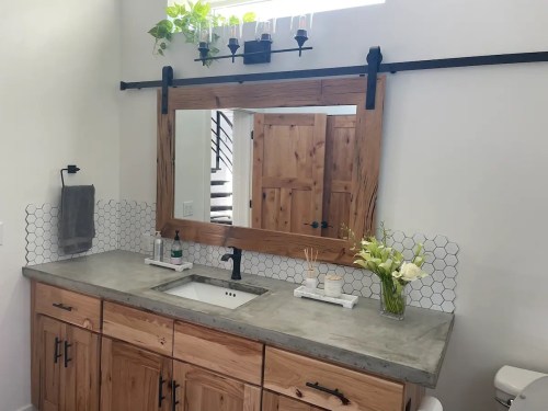 A modern bathroom with a wooden mirror, concrete sink, hexagonal tiles, and plants on a shelf above.