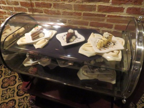 A glass display case showcasing various elegant desserts on white plates, set against a brick wall.