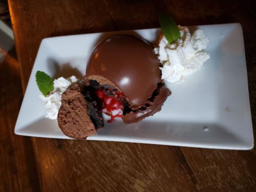 A chocolate dessert with a glossy shell, filled with cherry sauce, served with whipped cream and mint leaves on a plate.