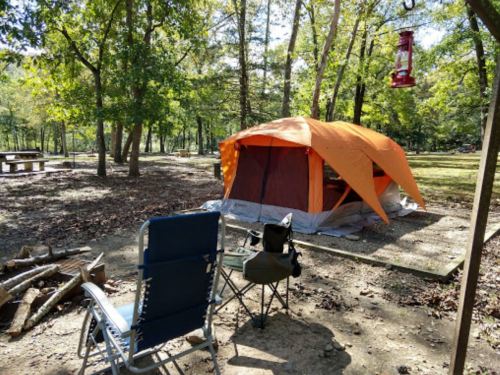 A bright orange tent set up in a wooded campsite, with camping chairs and a picnic table nearby.
