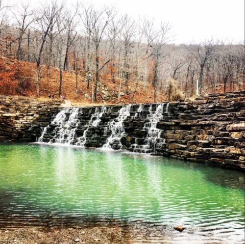 A serene waterfall cascades over stone into a calm, green pool, surrounded by bare trees and autumn foliage.