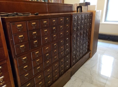 A wooden card catalog with multiple drawers, arranged in a grid, against a wall in a library setting.