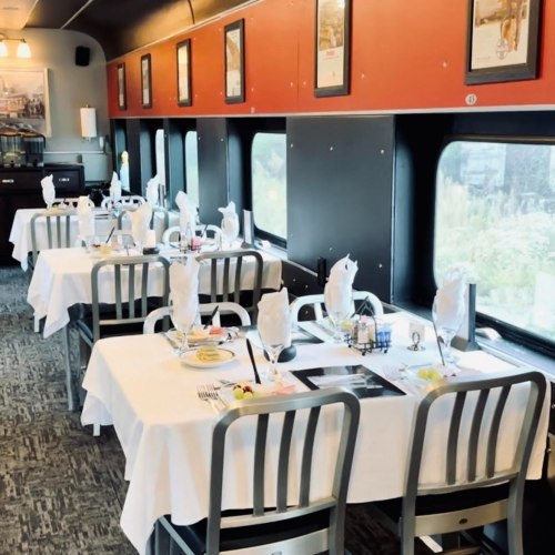 Interior of a dining car with neatly set tables, white tablecloths, and large windows showcasing a scenic view.