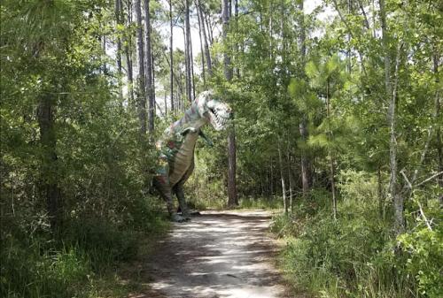 A colorful dinosaur statue stands beside a dirt path in a lush, green forest.