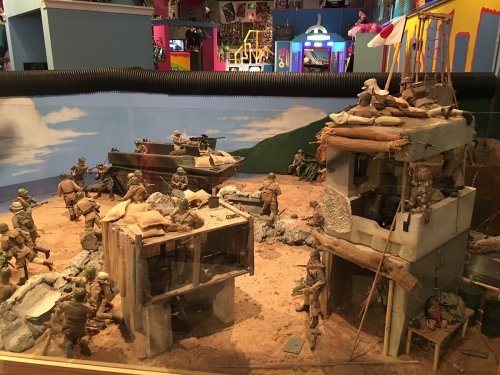 A detailed diorama depicting soldiers in a desert landscape with buildings, vehicles, and a painted backdrop.