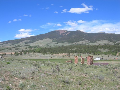 Scenic landscape featuring mountains, green hills, and remnants of brick structures under a blue sky with clouds.