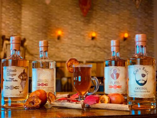 Three bottles of flavored spirits with a spiced drink and dried apple slices on a wooden table, set in a cozy bar.