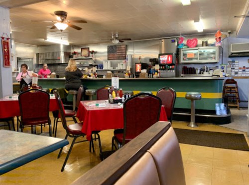 A cozy diner interior with red tablecloths, a counter, and patrons enjoying their meals. Decor includes an American flag.