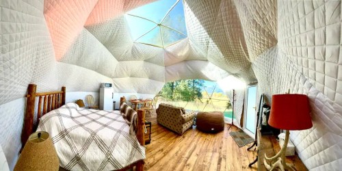 A cozy, uniquely designed interior of a geodesic dome with a bed, seating area, and large windows showcasing nature.
