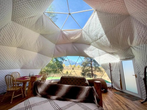 Interior of a geodesic dome with cozy furnishings, large windows, and a scenic view of nature outside.