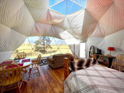 Interior of a geodesic dome with cozy furnishings, large windows, and a scenic view of nature outside.