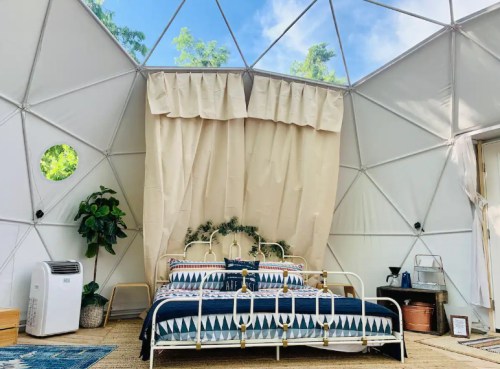 Cozy interior of a geodesic dome with a bed, plants, and natural light streaming through large windows.