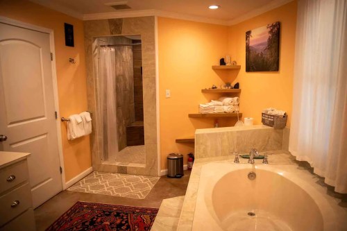 Bright bathroom featuring a shower, a large tub, neatly stacked towels, and warm orange walls.