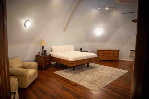 A spacious bedroom featuring a bed, nightstand, armchair, dresser, and soft lighting on blue walls.