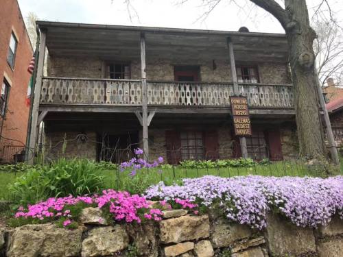 Historic stone house with a wooden balcony, surrounded by colorful flowers and greenery.