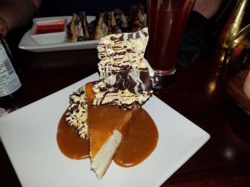 A slice of cheesecake topped with caramel, chocolate drizzle, and a decorative chocolate piece, served on a white plate.