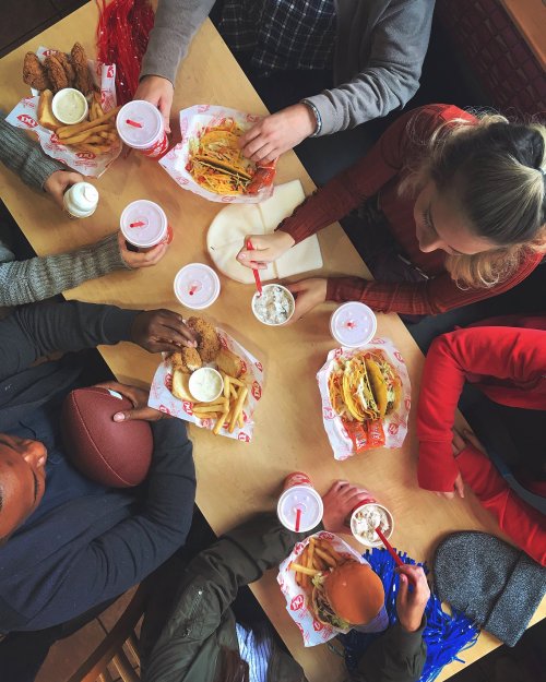 A group of people enjoying a meal with fried chicken, tacos, fries, and drinks at a table, celebrating together.