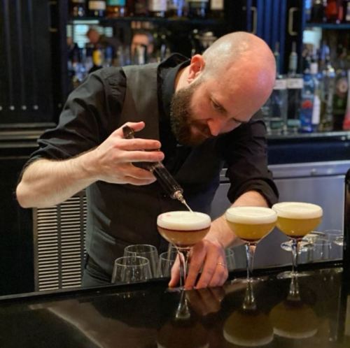 A bartender carefully pours a cocktail into a glass, with three finished drinks on the bar in front of him.