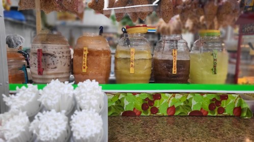 Colorful jars of various drinks labeled "Melon," "Piña," "Mango," and "Guava," with whipped cream cups in the foreground.