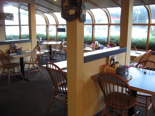 Empty restaurant interior with wooden chairs and tables, large windows, and a view of greenery outside.