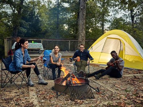 Four friends gather around a campfire, enjoying snacks and drinks near a yellow tent in a wooded area.