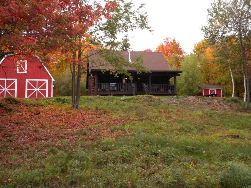 A cozy cabin surrounded by vibrant autumn foliage and a red barn in a scenic rural landscape.