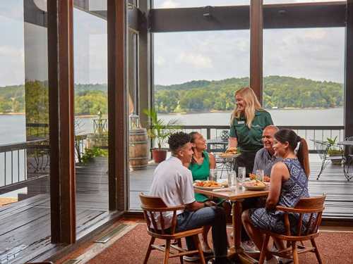 A group of four people dining at a table with a server, overlooking a scenic lake view through large windows.