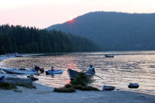 A serene lakeside scene at sunset, with boats on the water and mountains in the background.