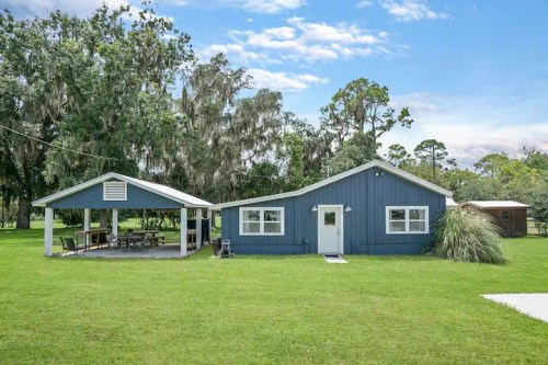A blue house with a porch and a separate covered area, surrounded by green grass and trees under a clear sky.