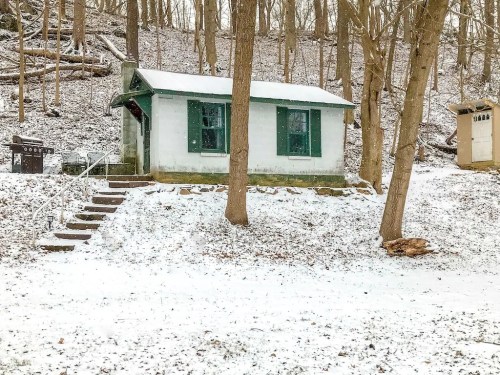 A small, white cabin with green shutters sits in a snowy landscape surrounded by bare trees.