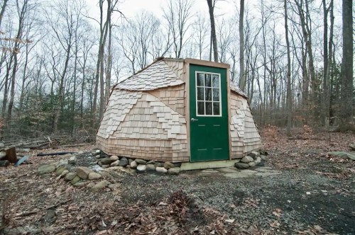 A small, round wooden cabin with a green door, surrounded by stones, set in a wooded area during winter.
