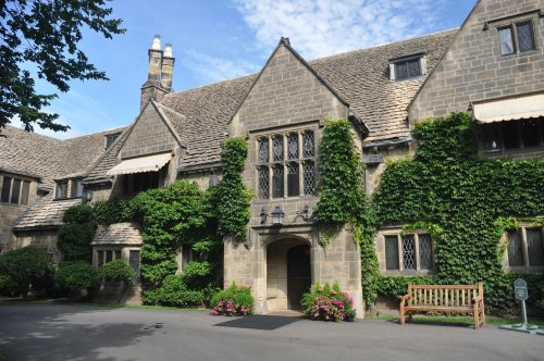 A historic stone building covered in greenery, featuring large windows and a welcoming entrance with a bench.
