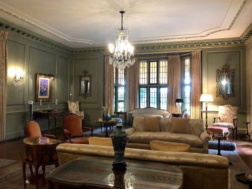 Elegant living room with green walls, vintage furniture, a chandelier, and large windows letting in natural light.