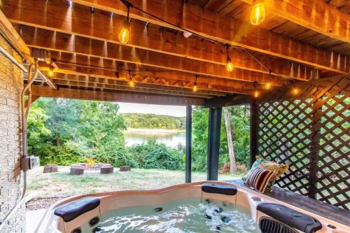 A hot tub under a wooden pergola with string lights, overlooking a lush green landscape and a body of water.