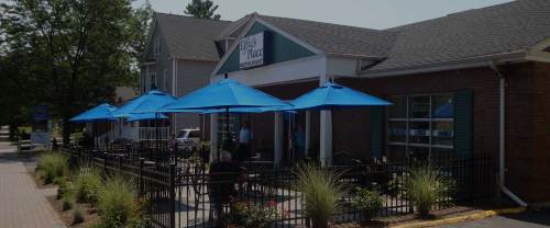 Exterior view of a restaurant with blue umbrellas on a patio, surrounded by greenery and a brick facade.