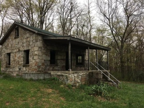 A stone house with a porch, surrounded by trees and greenery, on a cloudy day.