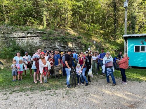 A large group of people, including children, gathered outdoors near a blue cabin and rocky hillside, smiling and posing for a photo.
