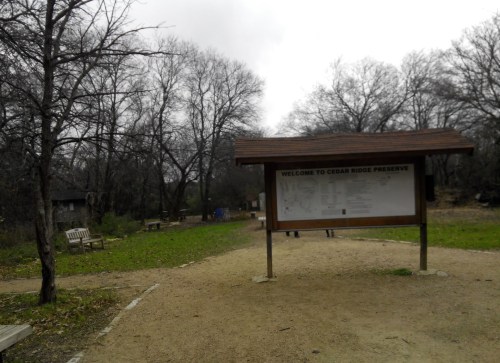 Sign welcoming visitors to Cedar Ridge Preserve, surrounded by bare trees and a gravel path. Overcast sky in the background.