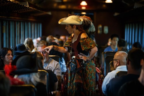 A woman in a cowboy hat and colorful dress interacts with passengers on a vintage train, surrounded by diverse travelers.