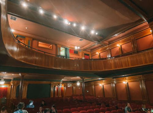 Interior of a vintage theater with wooden accents, red seating, and a balcony, softly lit with warm lights.