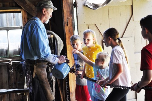 A blacksmith demonstrates a technique to a group of children in a workshop, engaging them with hands-on learning.