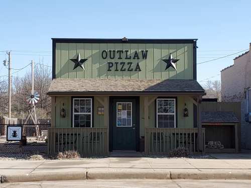 A green building with a sign reading "Outlaw Pizza," featuring stars and a rustic design, set against a clear blue sky.
