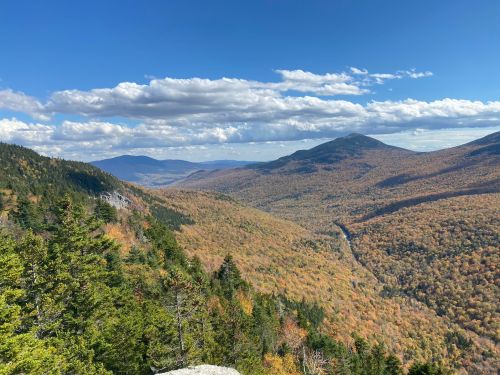 A panoramic view of a colorful autumn landscape with mountains and a valley under a blue sky with scattered clouds.