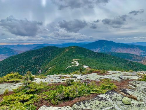 A panoramic view of green mountains under a cloudy sky, with rocky foreground and distant peaks in the background.