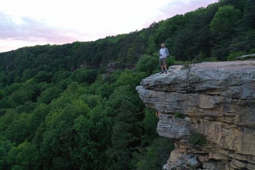 A person stands on a rocky cliff overlooking a lush green forest at sunset.